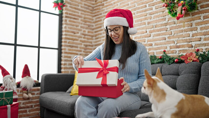 Young hispanic woman with chihuahua dog unpacking gift sitting on the sofa at home
