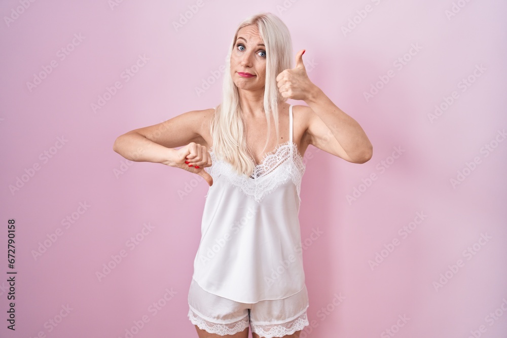 Sticker Caucasian woman wearing pajama wearing pink background doing thumbs up and down, disagreement and agreement expression. crazy conflict