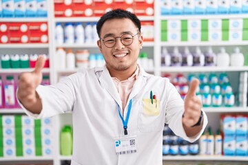 Chinese young man working at pharmacy drugstore looking at the camera smiling with open arms for hug. cheerful expression embracing happiness.