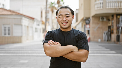 Cheerful young chinese man, radiating confidence and joy, spotted smiling and standing with crossed arms gesture on a bustling urban street