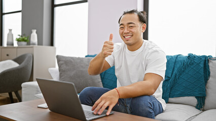 Cheerful young chinese guy happily sitting on home sofa, giving the thumbs up gesture while using a...