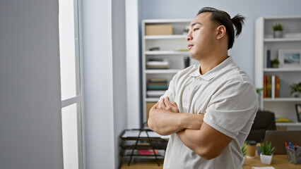 Young chinese man, a focused business worker, throws a side glance out the office window. standing, arms folded, he seems troubled indoors, contemplating a workplace problem.