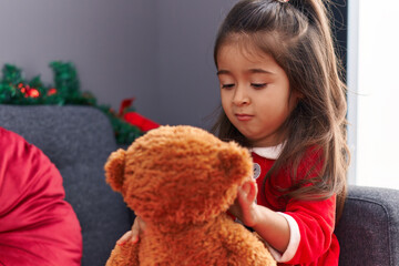 Adorable hispanic girl playing with teddy bear sitting on sofa by christmas decoration at home