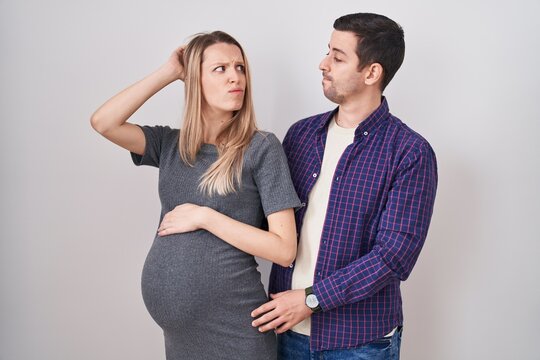 Young couple expecting a baby standing over white background confuse and wondering about question. uncertain with doubt, thinking with hand on head. pensive concept.