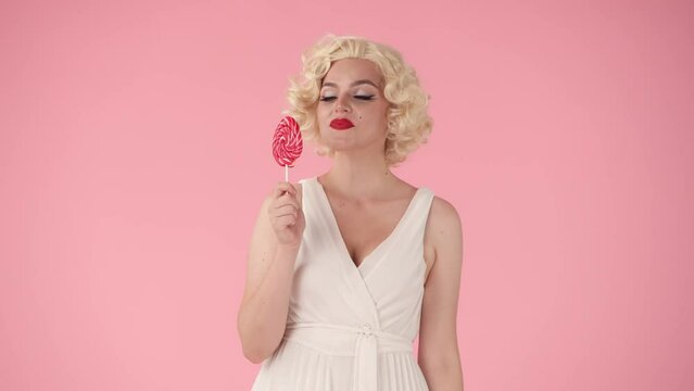 Woman, surprised says wow, showing a colorful lollipop. Woman happily licks the sweet candy on a stick, bites the lollipop with her teeth. Woman in the image of on pink background.