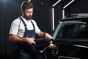 Side view on Young professional car service male worker with orbital polisher, polishing black...