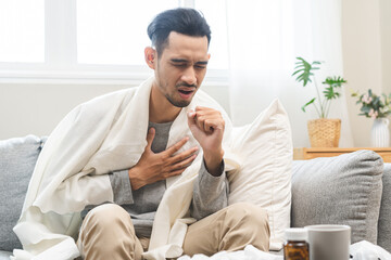 Sick, influenza concept, suffering asian young man under blanket have a fever, flu or sore throat and sneezing until chest pain while illness male sitting on sofa at home. Health care on virus person.