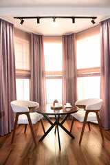 The room is cozy with curtains and a window with a table chairs and tea on the table