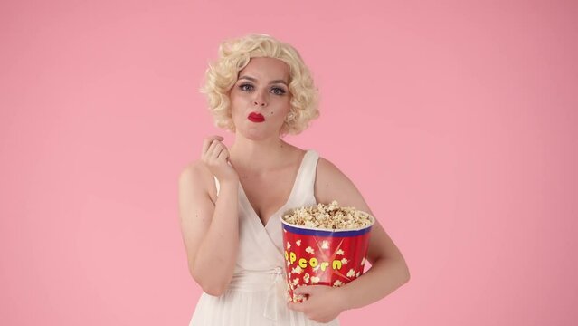 Portrait of a woman enjoying watching a movie and eating popcorn from a large bucket. The woman is laughing, watching a comedy. Woman in the image of in the studio on a pink background.