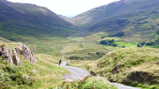 Rear view of bicycle rider group of four riding downhill mountain road towards Duddon Valley in Lake District, England, United Kingdom, one cyclist stops to take a picture, Hard Knott Pass viewpoint.