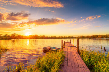 Sunset over the lake. Wooden village bridge with a boat. Cloudy sky. Ripples in the water. Reed thickets. View from the bridge. Russia, Europe. - obrazy, fototapety, plakaty