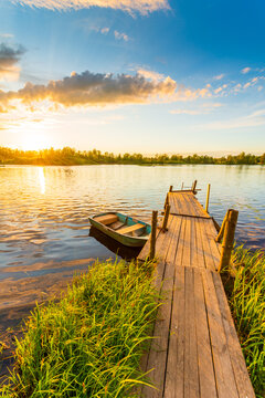 Sunset over the lake. Wooden village bridge with a boat. Cloudy sky. Ripples in the water. Reed thickets. View from the bridge. Russia, Europe. Vertical shot.