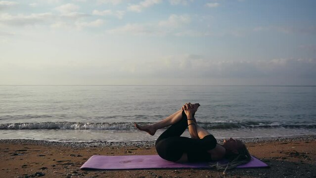 Slender woman doing yoga exercises on  seashore near the waves, dawn time, sun over  horizon