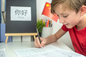 An 11 year old child is learning Chinese. a happy European boy sits at a desk, in front of him is a Chinese flag, on the desk is the hieroglyph “Hello” in Chinese, the boy is doing his homework  - obrazy, fototapety, plakaty