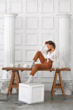 Young stylish woman in white blouse and beige pants sitting on wooden table in old greek decorated room. Full length portrait of modern woman among colomn