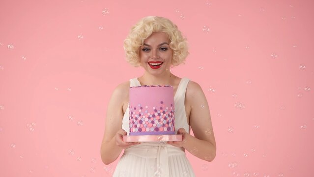 Joyful woman surrounded by soap bubbles holding a festive colorful cake. Woman celebrating birthday, victory, success. Woman in the image of in studio on pink background.