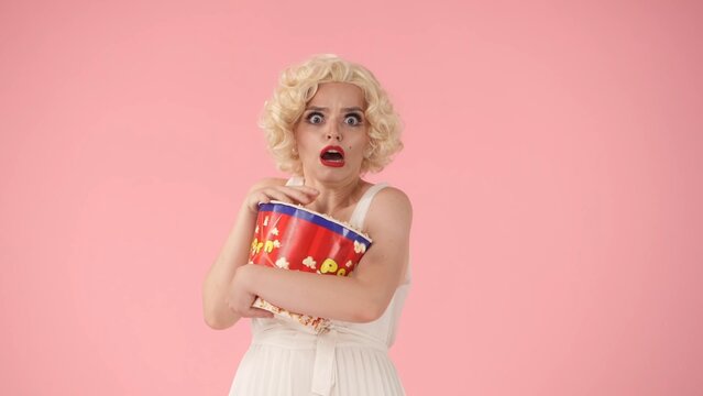 Portrait of frightened woman with big bucket of popcorn. Horror movie. Woman in the image of in studio on pink background.