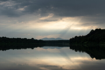 Landscape with mountain silhouette reflecting in river and overcast clouds with glowing sun rays