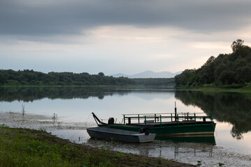 Obraz premium Landscape with moored boats in river against mountain silhouette in distance and cloudy sky