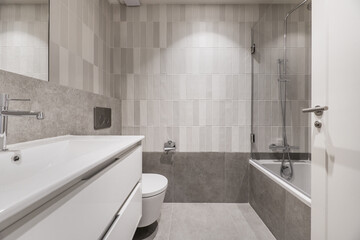 Newly installed small modern bathroom with bathtub with glass screen, multi-tone gray tiling, white...