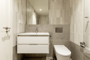 Frontal image of small modern bathroom with gray stone-like tiles, mirror on the wall, white wooden cabinet with drawers under the sink