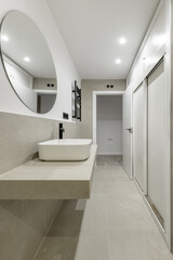 Small modern bathroom with gray stone imitation tiles, circular mirror on the wall, white porcelain sink, built-in wardrobe, towel drying radiator and black taps