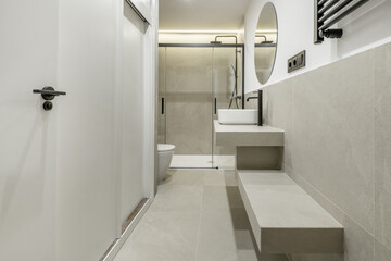 Small modern bathroom with gray stone imitation tiles, circular mirror on the wall, white porcelain sinks, built-in wardrobe and black faucets