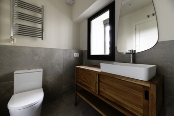 Small modern design bathroom with gray stone effect tiles, mirror on the wall, white porcelain sink cantilevered on wooden cabinet, towel drying radiator and chrome taps, black aluminum window