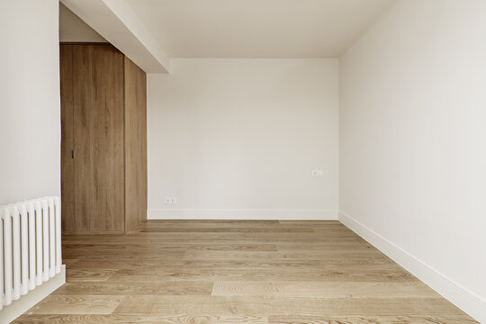 Empty room with white painted walls, wooden floors, wardrobe in a corner and radiator on a wall