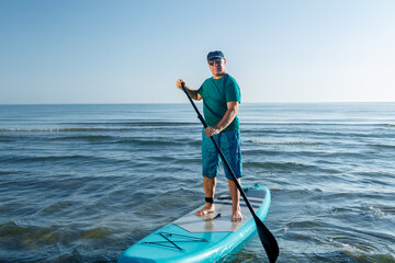 A middle-aged man in shorts and a T-shirt on a SUP board near the sea.
