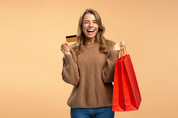 Beautiful smiling woman winks holding credit card and shopping bags isolated on beige background 