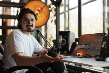 Male musician in white t shirt and eyeglasses sitting near laptop and microphone with crossed legs...