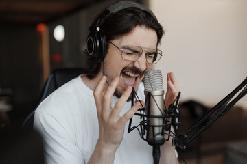Side view of passionate male singer in headphones sitting at table near microphone and recording...