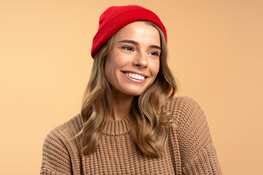 Portrait of beautiful smiling woman wearing red hipster hat and stylish winter sweater looking away isolated on beige background. Fashion, natural beauty concept  