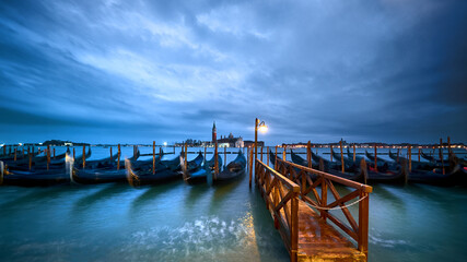 Italy, gondolas moored by Venice Embankment at night. Lonely light on wet wooden pier.