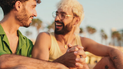 Romantic moments, homosexual couple enjoying communication with each other while sitting on the beach at sunrise, crossing their arms and looking at each other with love