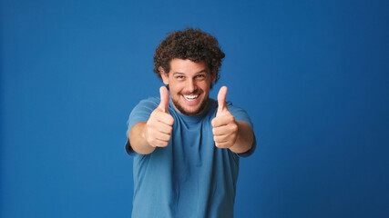 Happy guy with curly hair dressed in blue t-shirt looking at camera and showing double thumbs up isolated on blue background in studio