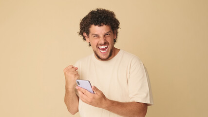 Happy guy with curly hair dressed in beige T-shirt, with mobile phone in his hands, celebrating victory, receiving good news while standing in studio on beige background in the studio