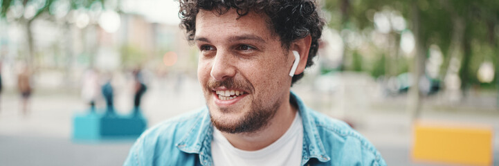 Close-up portrait of young bearded man in denim shirt sitting in wireless headphones and drinking...
