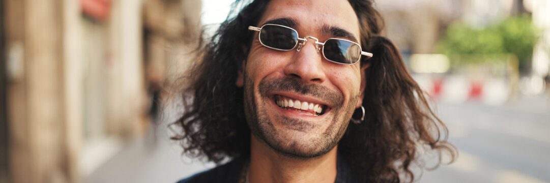 Young italian guy with long curly hair and stubble walks smiling down the street. Stylish man with sunglasses and lot of chains smiling and looking around