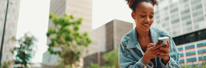 African girl with ponytail wearing denim jacket leaving voice message on mobile phone against modern city background. - obrazy, fototapety, plakaty