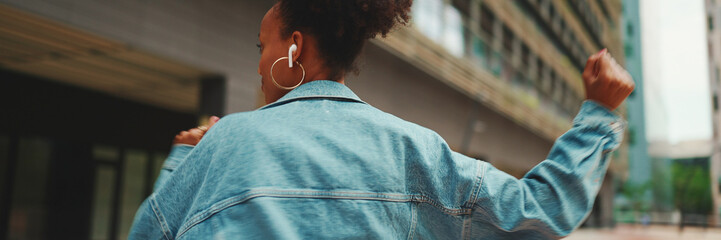 Closeup, smiling African girl with ponytail wearing denim jacket, in crop top with national pattern listening to music on headphones and dancing outdoors.