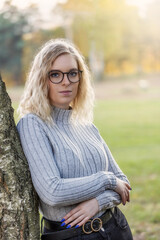 Obraz premium Autumn portrait of serious beautiful young woman in glasses posing leaning against a tree. Vertically.