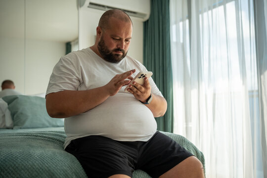 Fat bearded bald guy with smartphone sits on hotel bed, dials number on phone, types message, scrolls social networks, watches news, weather, browses web pages. Obese man with mobile phone chatting