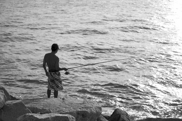 High angle grayscale shot of a male fishing on a rocky shoreline