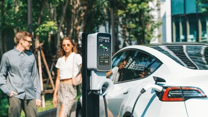 Fotobehang Young couple travel with EV electric car charging in green sustainable city outdoor garden in summer shows urban sustainability lifestyle by green clean rechargeable energy of electric vehicle innards © Summit Art Creations