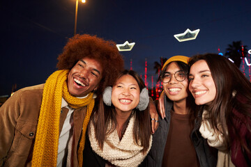Four joyful friends posing smiling selfie mobile looking at camera standing hugging at christmas winter night outdoor. Multi-ethnic group of cheerful people in amusement park having fun together. - obrazy, fototapety, plakaty