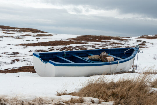 A white wooden traditional Rodney boat or small fishing vessel with blue trim and a rib down the middle in a grassy field. The rowing boat has a white coat of paint and there's snow on the ground.