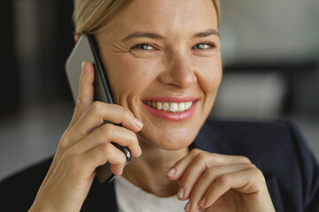 Cheerful businesswoman talking phone sitting on modern office background and looks camera