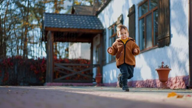 Joyful Caucasian toddler running by the yard in autumn. Energetic and active walk outdoors on sunny day.
